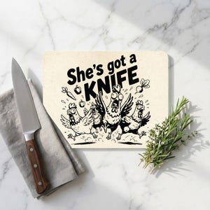May include: A rectangular chopping board with the text "She's got a KNIFE" and a cartoon of chickens running away. A stainless steel knife with a wooden handle and a sprig of rosemary are also present.
