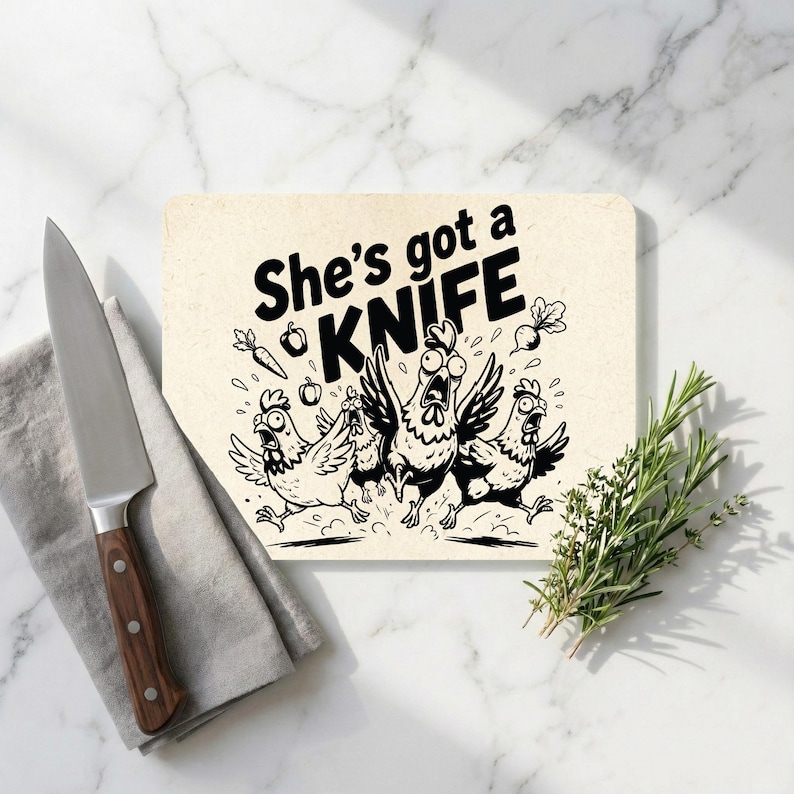 May include: A rectangular chopping board with the text "She's got a KNIFE" and a cartoon of chickens running away. A stainless steel knife with a wooden handle and a sprig of rosemary are also present.