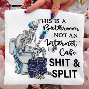 May include: White t-shirt with a humorous graphic of a skeleton on a toilet, holding a phone. The text reads "THIS IS A BATHROOM NOT AN INTERNET CAFE SHIT & SPLIT". The skeleton is wearing blue jeans.