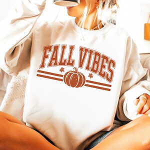 May include: A cream-colored crewneck sweatshirt featuring the text FALL VIBES in a distressed rust-orange font. Below the text is a graphic of a pumpkin flanked by two small leaves, all centered between two horizontal lines in the same rust-orange color.