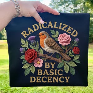 May include: A navy blue square textile with the words "RADICALIZED BY BASIC DECENCY" in gold lettering. The design features a crocheted robin perched on a branch with crocheted roses, flowers, and leaves.