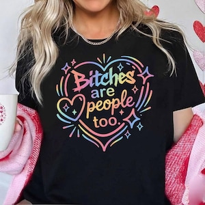 Puede incluir: Camiseta negra con la frase "Bitches are people too" en un diseño de corazón arcoíris. El gráfico incluye estrellas y líneas. Una camiseta informal para el uso diario.
