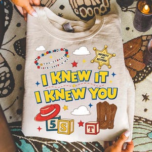 May include: A beige crewneck sweatshirt featuring a graphic print with the text I KNEW IT I KNEW YOU, a sheriff badge with INFINITY AND BEYOND, a beaded bracelet with YOU'VE GOT A FRIEND IN ME, a red cowboy hat, brown cowboy boots, and alphabet blocks.