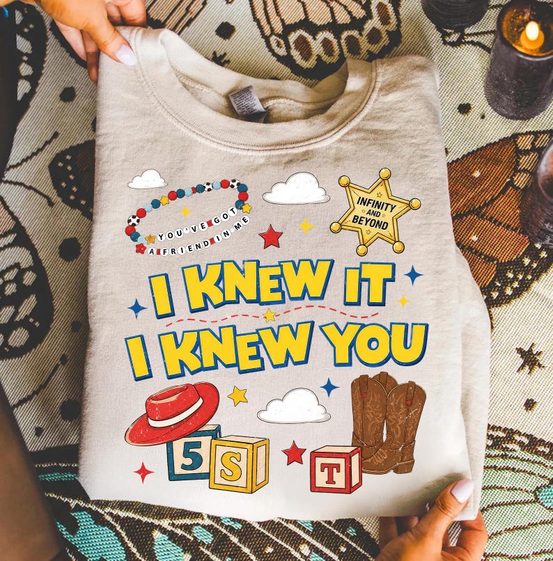 May include: A beige crewneck sweatshirt featuring a graphic print with the text I KNEW IT I KNEW YOU, a sheriff badge with INFINITY AND BEYOND, a beaded bracelet with YOU'VE GOT A FRIEND IN ME, a red cowboy hat, brown cowboy boots, and alphabet blocks.