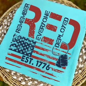 K&ouml;nnte beinhalten: Hellblaues T-Shirt mit einem verwitterten Design der amerikanischen Flagge in Rot und Marineblau. Das Shirt zeigt den Text "REMEMBER EVERYONE DEPLOYED RED EST. 1776" und eine Hundemarken-Grafik.