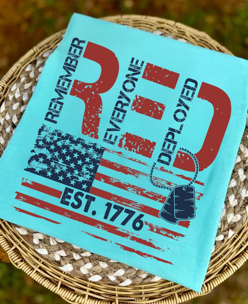 K&ouml;nnte beinhalten: Hellblaues T-Shirt mit einem verwitterten Design der amerikanischen Flagge in Rot und Marineblau. Das Shirt zeigt den Text "REMEMBER EVERYONE DEPLOYED RED EST. 1776" und eine Hundemarken-Grafik.