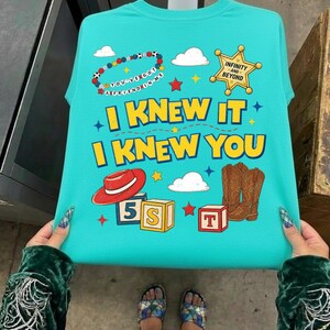May include: A bright turquoise t-shirt featuring a graphic print with the text "I KNEW IT I KNEW YOU" in yellow. The design includes a beaded bracelet, a gold sheriff star badge, a red cowboy hat, brown cowboy boots, and alphabet blocks.