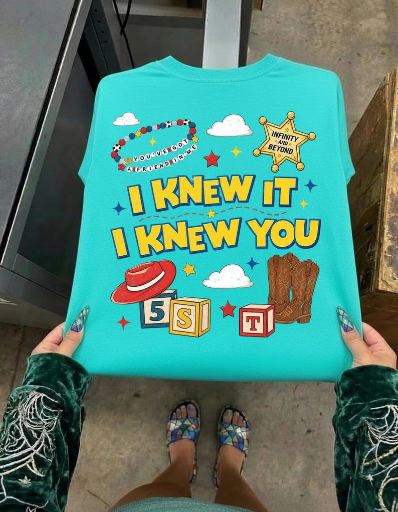 May include: A bright turquoise t-shirt featuring a graphic print with the text "I KNEW IT I KNEW YOU" in yellow. The design includes a beaded bracelet, a gold sheriff star badge, a red cowboy hat, brown cowboy boots, and alphabet blocks.