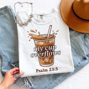 May include: White t-shirt featuring a graphic of an iced coffee drink with the text "my cup overflows" and "Psalm 23:5". The shirt has a crew neck and short sleeves. A silver necklace and a brown hat are also visible.