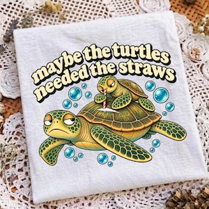 May include: A white t-shirt featuring a graphic of two green sea turtles with a red and white striped straw and a white line on the shell. The text above the turtles reads, maybe the turtles needed the straws, in a bold, dark, retro-style font.