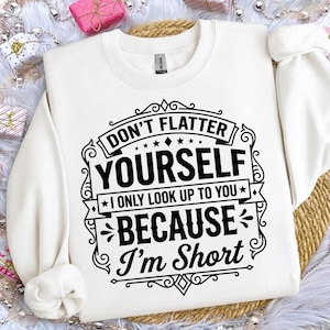 May include: White sweatshirt with the text "DON'T FLATTER YOURSELF, I ONLY LOOK UP TO YOU BECAUSE I'M SHORT" in a black decorative font. The sweatshirt has a crew neck and long sleeves.