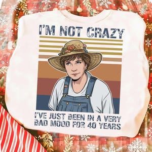 Può includere: Felpa bianca con la scritta "I'M NOT CRAZY" sopra una grafica di una donna con cappello di paglia e tuta. Il testo "I'VE JUST BEEN IN A VERY BAD MOOD FOR 40 YEARS" è sotto.