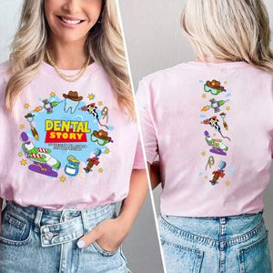 以下が含まれることがあります： 「DENTAL STORY」および「TO INFINITY AND BEYOND FOR OUR PATIENTS」というテキストがプリントされたライトピンクのTシャツ。歯、歯科用チェア、さまざまな歯科用器具のカラフルなイラストがデザインされています。