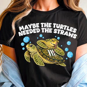May include: A black t-shirt featuring a graphic of two green sea turtles with blue bubbles. One turtle holds a red and white striped straw over three white lines on the other turtle's shell. The text reads: MAYBE THE TURTLES NEEDED THE STRAWS.