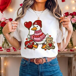 May include: White t-shirt featuring a cartoon duck wearing a red hat and sweater, holding a heart-shaped balloon. A teddy bear with a heart and a heart-shaped gift box are also shown. The design has pink and red accents, perfect for Valentine's Day.
