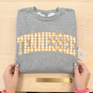 Pode incluir: Uma camisola cinzenta mesclada de gola redonda com a palavra TENNESSEE num padrão de xadrez vichy laranja e branco arqueado. Uma régua de metal dourado está colocada horizontalmente abaixo do texto, mostrando um comprimento de 15 centímetros.