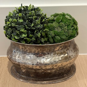 May include: A hammered silver-toned metal planter with a wide, shallow bowl shape. The planter contains two artificial green plant spheres. The planter sits on a wooden surface against a white wall.