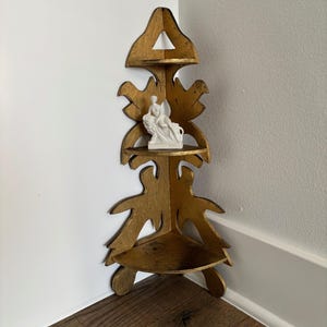 May include: A gold-painted wooden corner shelf with three tiers, featuring a decorative leaf design. A small white figurine sits on the middle shelf. The shelf is positioned in a corner against white and gray walls, with a wooden floor.