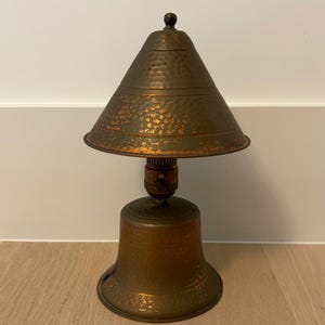 May include: A vintage copper desk lamp with a bell-shaped base and a conical shade. The lamp has a hammered texture and a warm, aged patina. The lamp is a decorative lighting fixture.