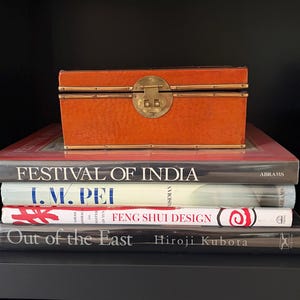 May include: An orange rectangular box with a brass clasp sits atop a stack of books. The books include titles like "FESTIVAL OF INDIA", "I.M. PEI", and "FENG SHUI DESIGN". The books are stacked on a black shelf.
