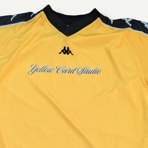 May include: A yellow long-sleeve shirt with black and light blue accents. The shirt features the text "Yellow Card Studio" and the Kappa logo. The text "COLLECTION" is at the top. The text "@thats.a.yellow" is at the bottom.