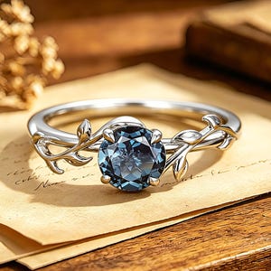 May include: A silver-toned ring featuring a round, deep blue faceted gemstone set in the center. The band is designed with delicate, sculpted metal leaves and vines extending from the setting, wrapping around the finger in a botanical motif.