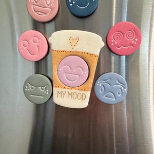 May include: A central cream-colored coffee cup magnet with the text MY MOOD and a heart icon, surrounded by six circular magnets in various colors including pink, blue, and green, each featuring a different embossed facial expression.