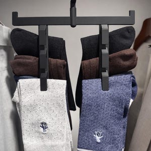 May include: A black sock organizer hanging in a closet, holding several pairs of socks. The socks are in various colors, including black, brown, gray, and blue. The organizer is suspended from a wooden rod.