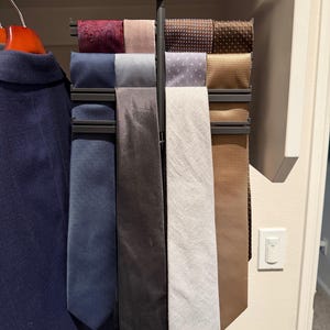 May include: A black plastic tie rack hanging from a wooden closet rod, holding eight neckties in various colors and textures including blue, grey, white, tan, brown, and patterned fabrics, organized in two vertical columns.