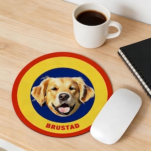 May include: A round mousepad featuring a smiling golden retriever dog. The dog is centered on a blue circle, surrounded by a yellow ring, and a red outer border. The word "BRUSTAD" is printed in red at the bottom. A white computer mouse, a coffee mug, and a notebook are also visible.