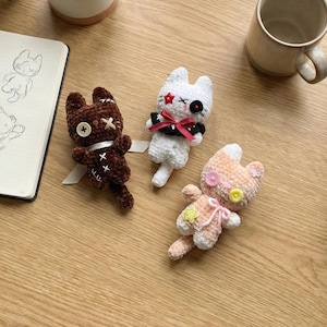 May include: Three handmade crochet cat dolls in brown, white, and peach colors. The brown cat has a button eye and stitched details. The white cat has a red star and black bow. The peach cat has a pink bow and button details. A notebook and mug are also visible.