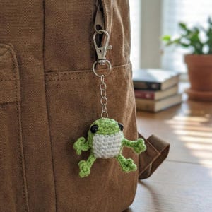May include: A green and white crocheted frog keyring attached to a brown rucksack. The frog has black eyes and is suspended by a silver chain and clasp. The rucksack is made of a textured fabric.