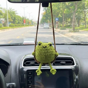 May include: A green crocheted frog car charm hanging from the rearview mirror. The frog is sitting on a brown swing, with its legs dangling. The car's dashboard is visible in the foreground, with a road and trees in the background.