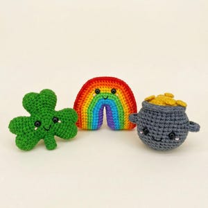 May include: Crocheted St. Patrick's Day themed items. A green shamrock, a rainbow with red, orange, yellow, green, blue, and purple stripes, and a gray pot filled with gold coins, all with smiling faces.