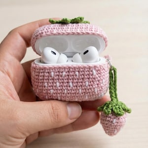 Könnte beinhalten: Ein rosa gehäkeltes AirPod-Etui in Erdbeerform mit weißen Samendetails und einem grünen Stiel und Blättern. Das Etui ist geöffnet und zeigt weiße AirPods. Ein kleiner Erdbeeranhänger hängt daran.