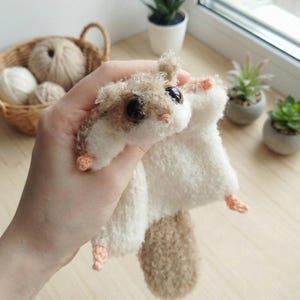 May include: A handmade plush sugar glider toy. The toy is held in a hand and features a fluffy, cream-colored body with brown and cream-colored accents on the head and tail. The sugar glider has large, dark eyes and small, peach-colored feet.