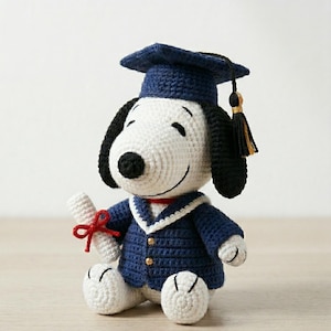 以下が含まれることがあります： ネイビーブルーの卒業帽子とガウンを着た、かぎ針編みのスヌーピー人形。人形は赤いリボンで結ばれた丸められた卒業証書を持っています。人形は黒い耳、黒い鼻、白い体を持っています。