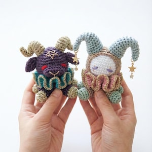 May include: Two handmade crochet dolls. One is purple with horns and a star, the other is beige with blue ears. Both have ruffled collars and star charms. The dolls are held in front of a white background.
