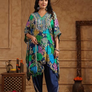 May include: A woman wearing a floral print kaftan top with matching navy blue pants. The kaftan features a V-neckline with embellishments and a coordinating purse. The outfit is adorned with colorful floral patterns in shades of blue, green, and purple.