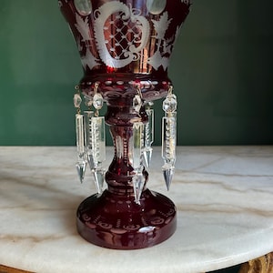 May include: A deep red glass goblet featuring intricate white etched patterns and a scalloped rim. The stem is adorned with multiple clear, faceted glass crystal prisms hanging from small metal loops, creating a decorative light-reflecting effect.