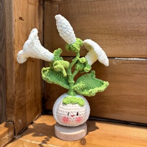 May include: A crocheted plant figurine featuring a round white base with a smiling face, pink cheeks, and green leaves. Three white trumpet-shaped flowers extend from the top on green stems, all mounted on a small, circular wooden stand.