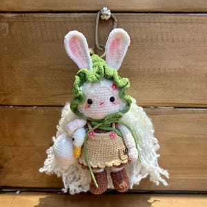 Könnte beinhalten: Handgemachte Häkelkaninchenpuppe mit grüner Haube, die eine weiße Gans hält. Das Kaninchen hat rosa gefütterte Ohren, ein beiges Kleid und braune Stiefel. Die Puppe hat einen weißen, flauschigen Schal und ist an einem silbernen Verschluss befestigt.