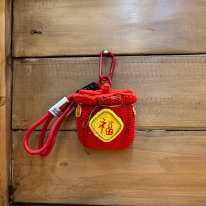 Puede incluir: Pequeño bolso rojo tejido a ganchillo con cierre de cordón y un emblema amarillo y dorado con caracteres chinos. El bolso está sujeto a un cordón rojo con un mosquetón rosa y un cierre dorado.