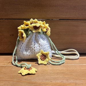 Puede incluir: Una bolsa blanca de ganchillo con cordón adornada con detalles de girasoles amarillos y marrones. La bolsa tiene un cordón verde y una correa crema. Adornos de girasol adicionales están dispersos alrededor de la bolsa.