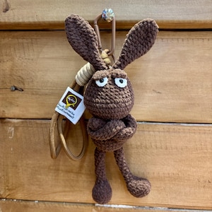 May include: A brown crocheted rabbit keychain with long ears, white eyes with black pupils, and arms crossed over its chest. The figure is attached to a tan lanyard and a brown plastic carabiner, featuring a small white tag with yellow text.