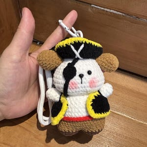 May include: A crocheted plush keychain featuring a white character dressed as a pirate. The figure wears a black and yellow hat with a white cross, a black eye patch, a black and yellow vest, and brown pants. It has pink cheeks and a white lanyard.