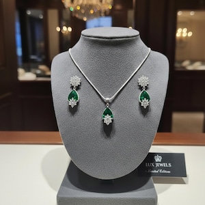 May include: A silver necklace and earring set displayed on a gray bust. The set features a teardrop-shaped, emerald-green gemstone pendant and matching earrings, each accented with a starburst of clear stones. The jewelry is presented on a gray display stand.