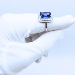 May include: A silver ring featuring a large, rectangular, deep blue gemstone, surrounded by a halo of smaller, clear stones. The ring is held up by a white gloved hand against a white background. The ring's band is thin and polished.