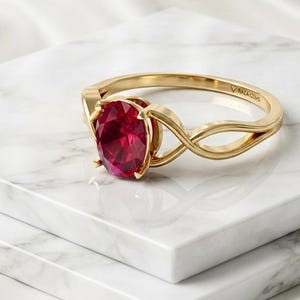 Könnte beinhalten: Ein Goldring mit einem oval geschliffenen, tiefroten Edelstein in einer Vier-Krappen-Fassung. Die Ringschiene hat ein geteiltes, gedrehtes Design und ist auf der Innenseite mit dem Text RAZA GEMS graviert. Das Metall hat eine polierte Oberfläche.