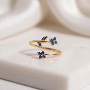 Könnte beinhalten: Ein offener Goldring im Zweig-Design mit zwei kleinen blauen Saphirblüten-Clustern und zwei marquise-geschliffenen blauen Saphirblättern. Die Ringschiene ist dünn und poliert und zeigt ein zartes botanisches Arrangement aus Edelsteinen.